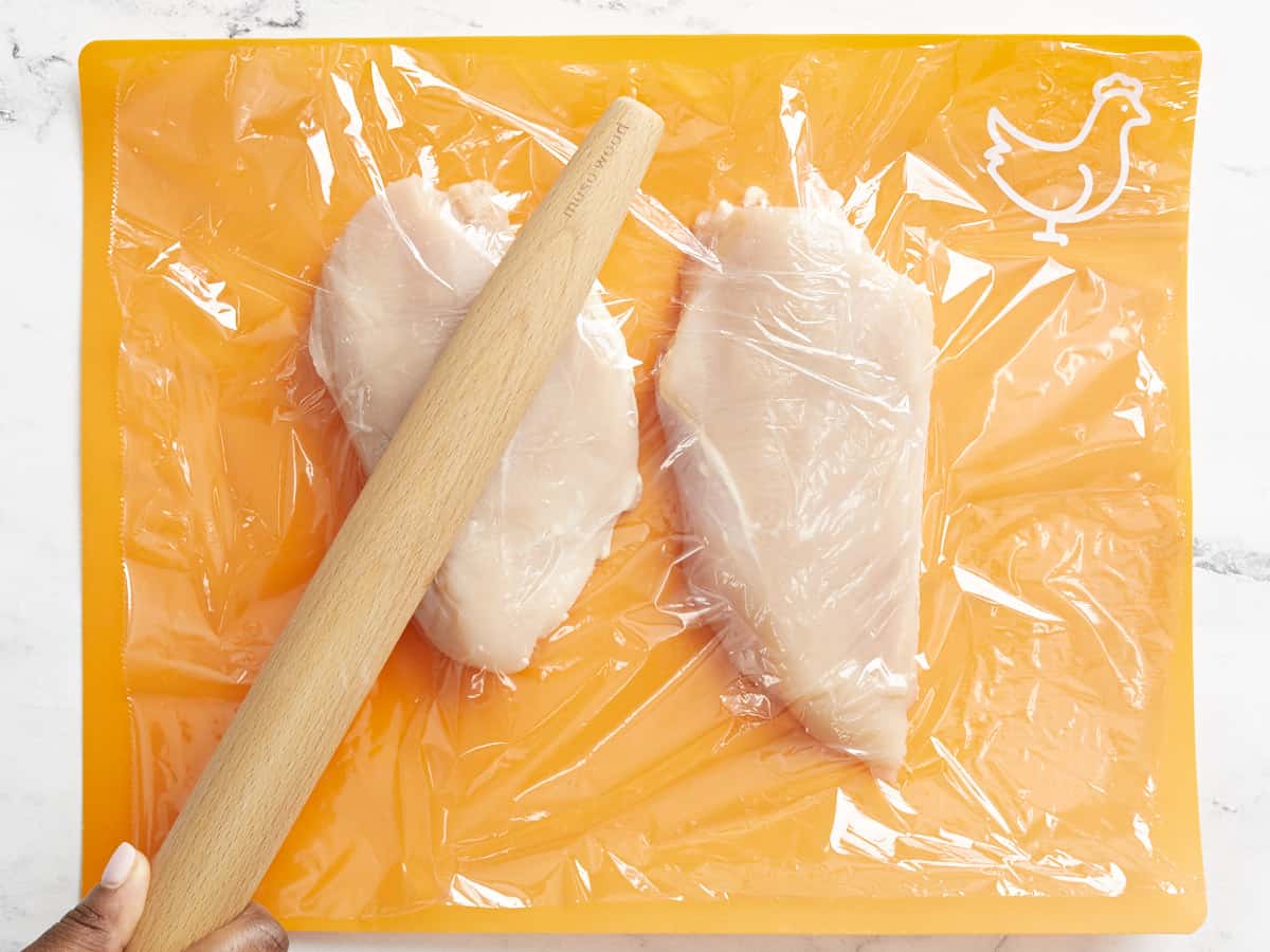 chicken breasts on a cutting board being pounded with a rolling pin.