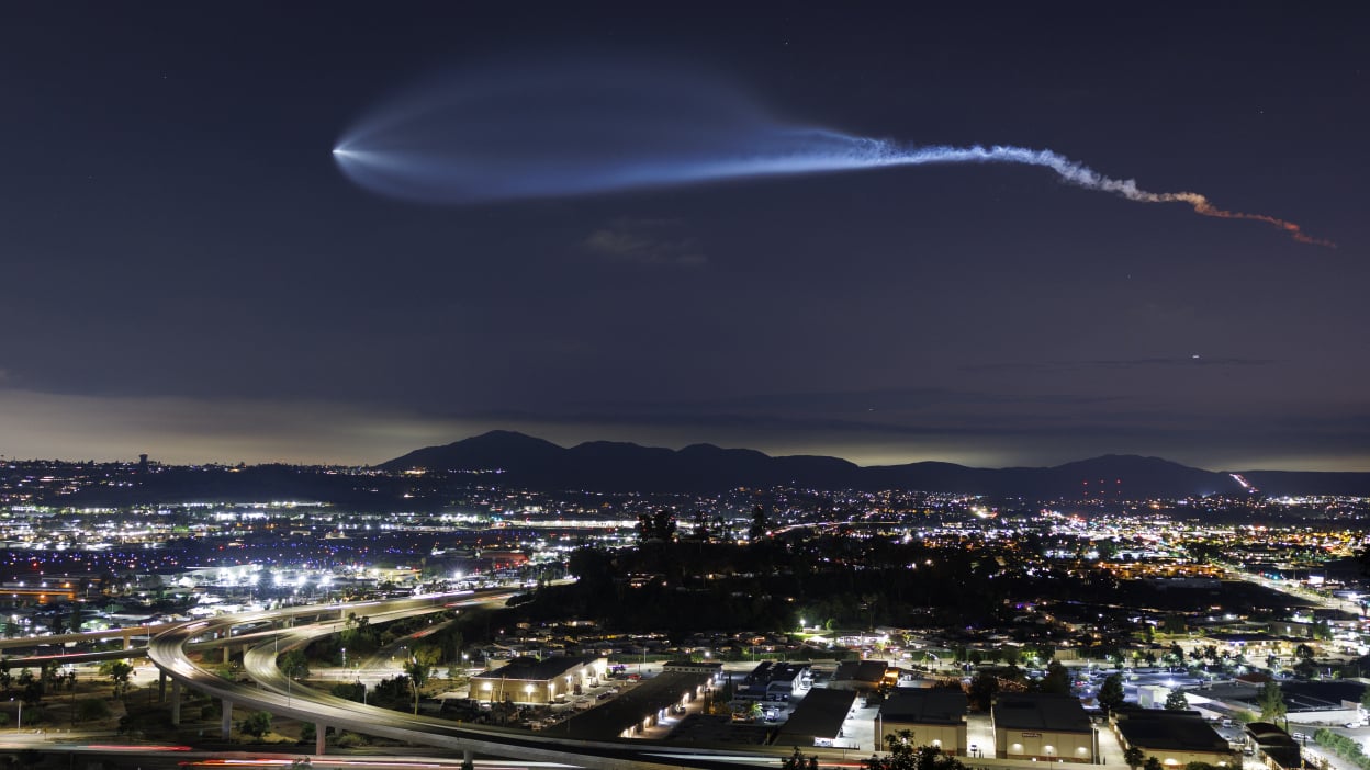 A SpaceX rocket flying through the night sky over a city skyline.