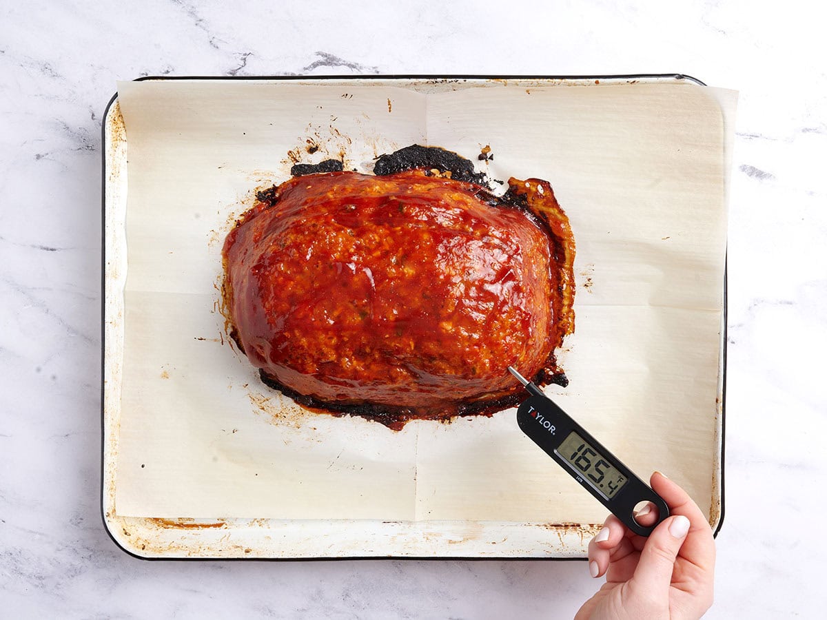 A thermometer reading 165°F being stuck into the chicken meatloaf on a white baking sheet lined with parchment paper.