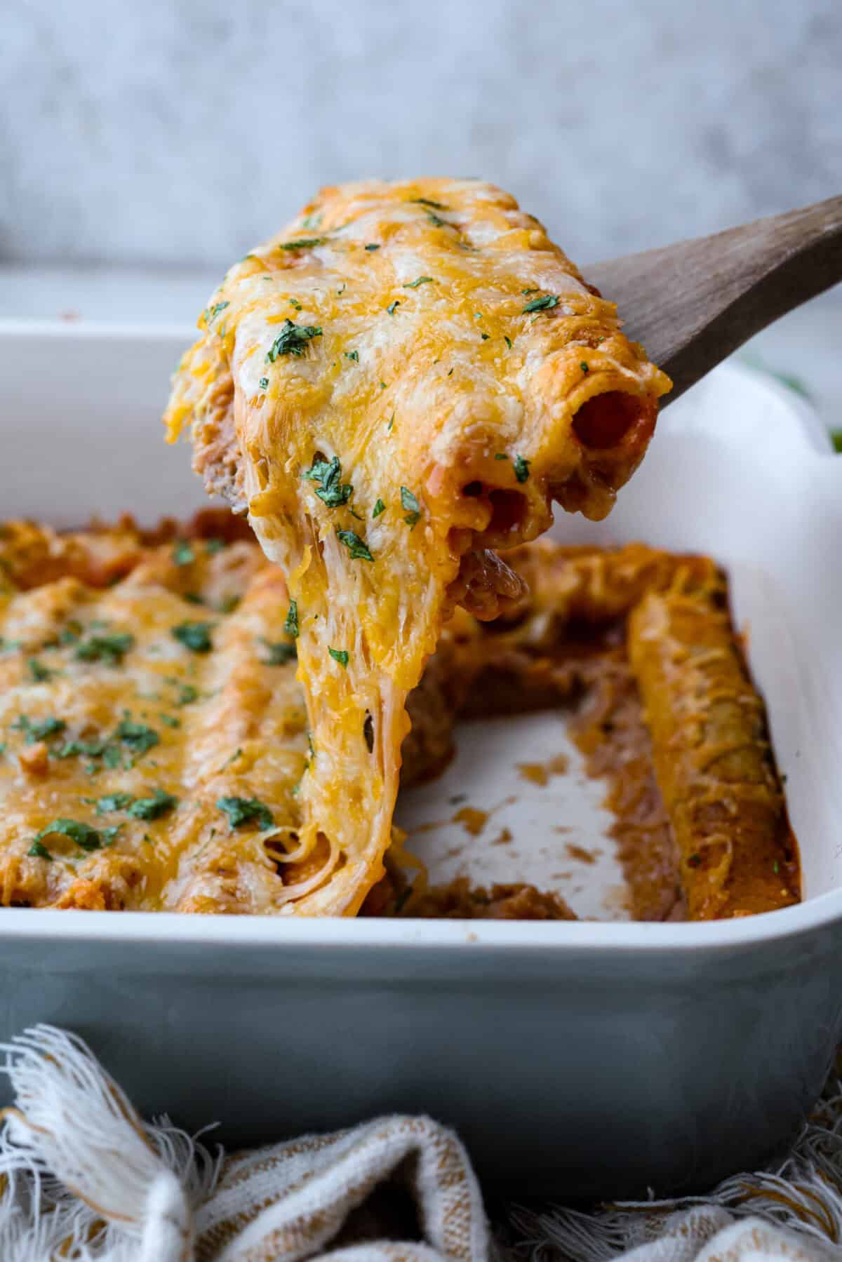 When you want enchiladas without the work, these Lazy Enchiladas are it! Four ingredients. Less than 30 minutes. Just layer frozen taquitos with beans, sauce, and cheese, bake until bubbly, and boom, dinner is served! Someone lifting some lazy enchiladas out of the pan with cheese stretching between.