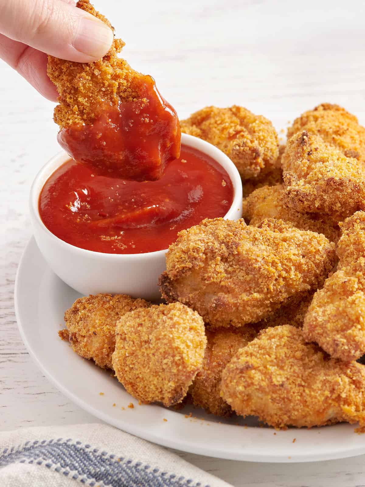 Homemade baked chicken nuggets are crispy on the outside, tender and juicy on the inside, easy to make, and make a delicious meal or snack. Budgetbytes.com Side view of a plate of homemade chicken nuggets with a hand dipping a nugget into a pot of ketchup.