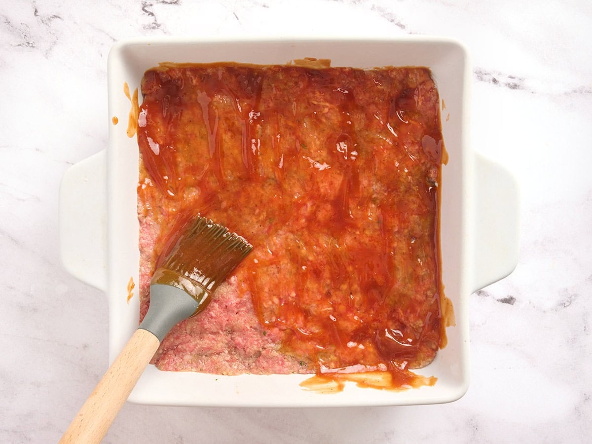 A ketchup glaze being spread across a meatloaf in a casserole dish.