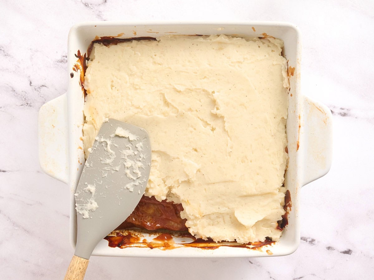 Mashed potatoes being spread over meatloaf casserole in a baking dish.