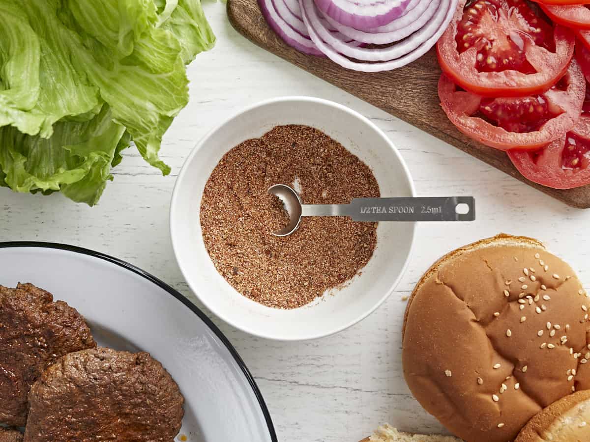 A bowl of burger seasoning surrounded by cooked burger patties and toppings.