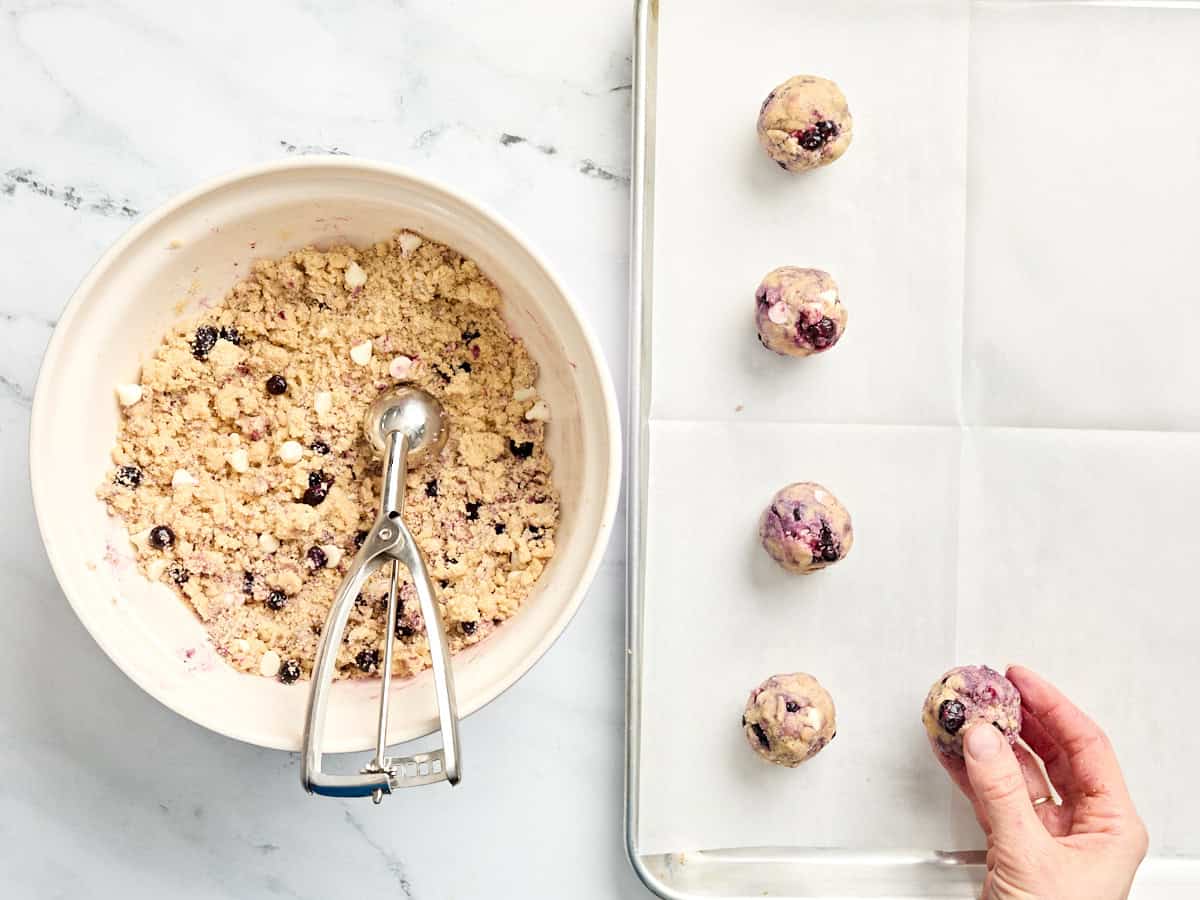 A bowl of blueberry cookie dough in a bowl with a scoop, next to the balls of cookie dough on a baking sheet.