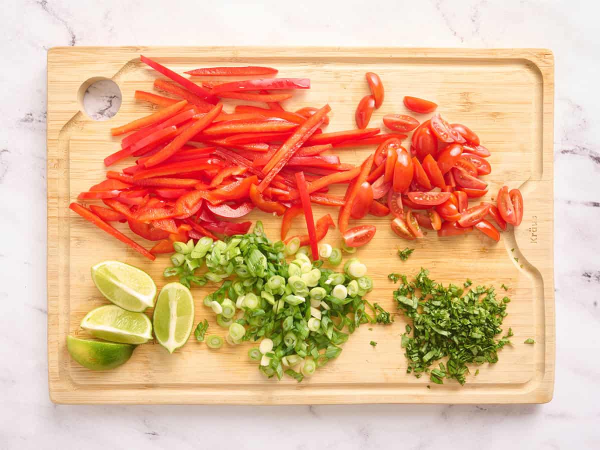 Sliced red bell pepper, sliced green onion, quartered lime wedges, sliced grape tomatoes and minced basil on a wooden cutting board.