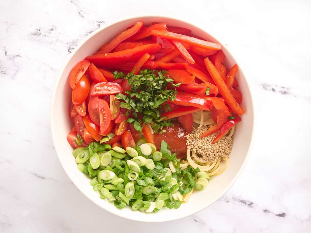 Sliced red bell pepper, sliced green onions, minced basil, sliced grape tomatoes and spaghetti in a bowl.
