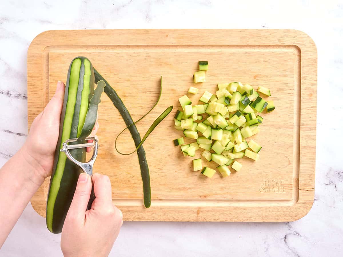 Hands peeling a cucumber, next to a wooden cutting board with diced cucumber on it.
