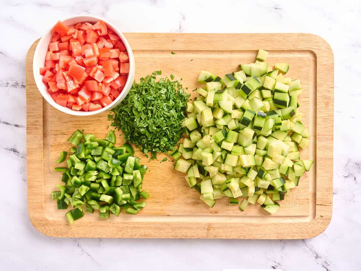 Chopped tomatoes, green bell pepper, cucumber, and parsley on a wooden cutting board.