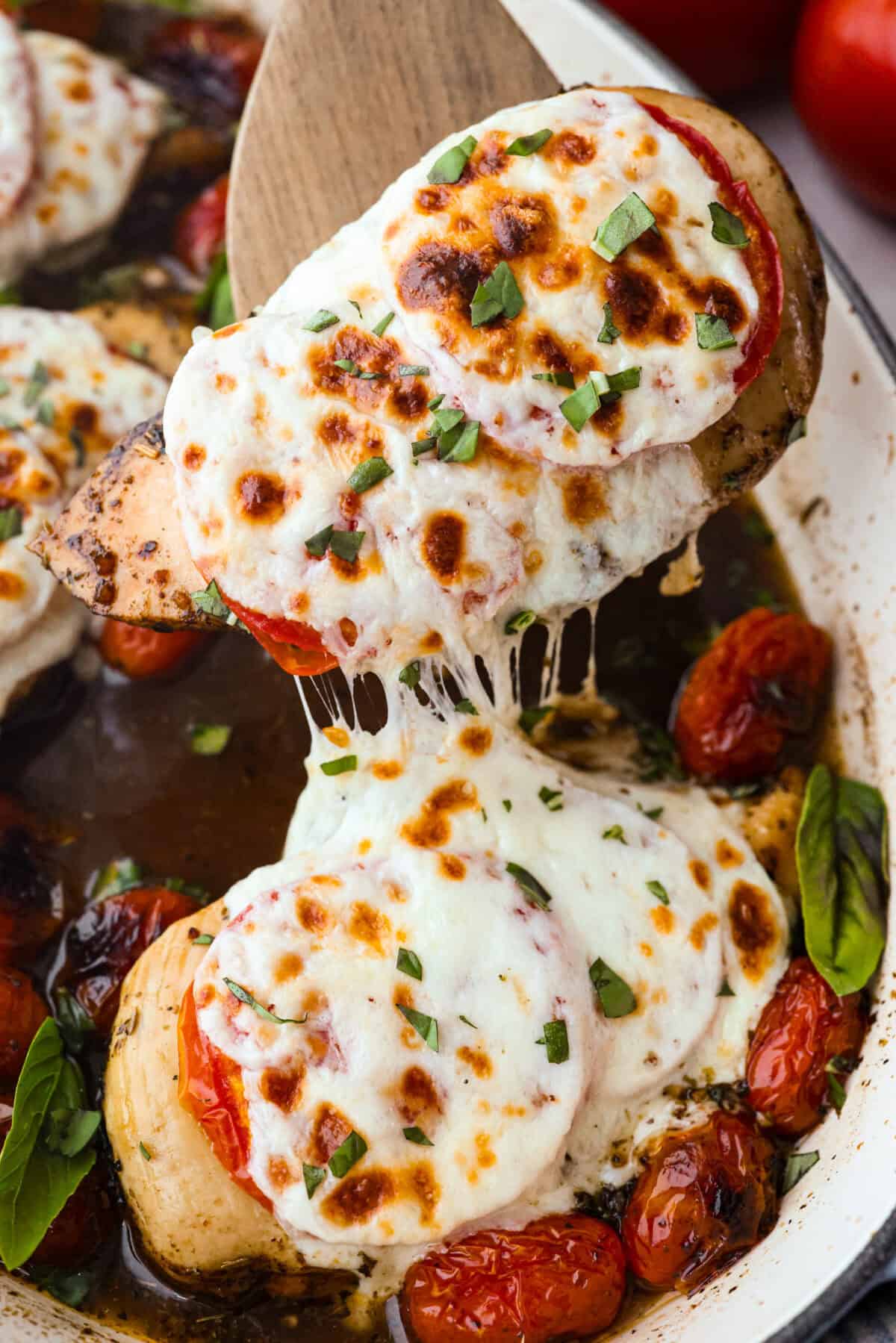 Juicy chicken, melty mozzarella, tomatoes, basil, and balsamic glaze all baked in one pan. Easy, fresh, and so good. Someone lifting the baked caprese chicken out of the pan with a wooden spatula.